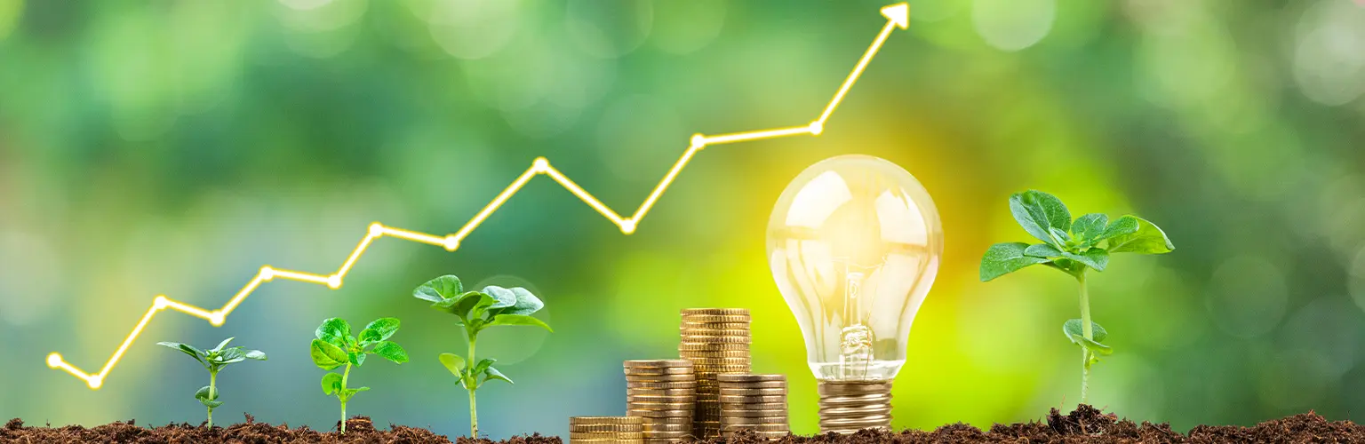 Close up view of a financial growth graph, coin stacks, plant pods and a glowing light bulb on soil shot against defocused lush foliage green background